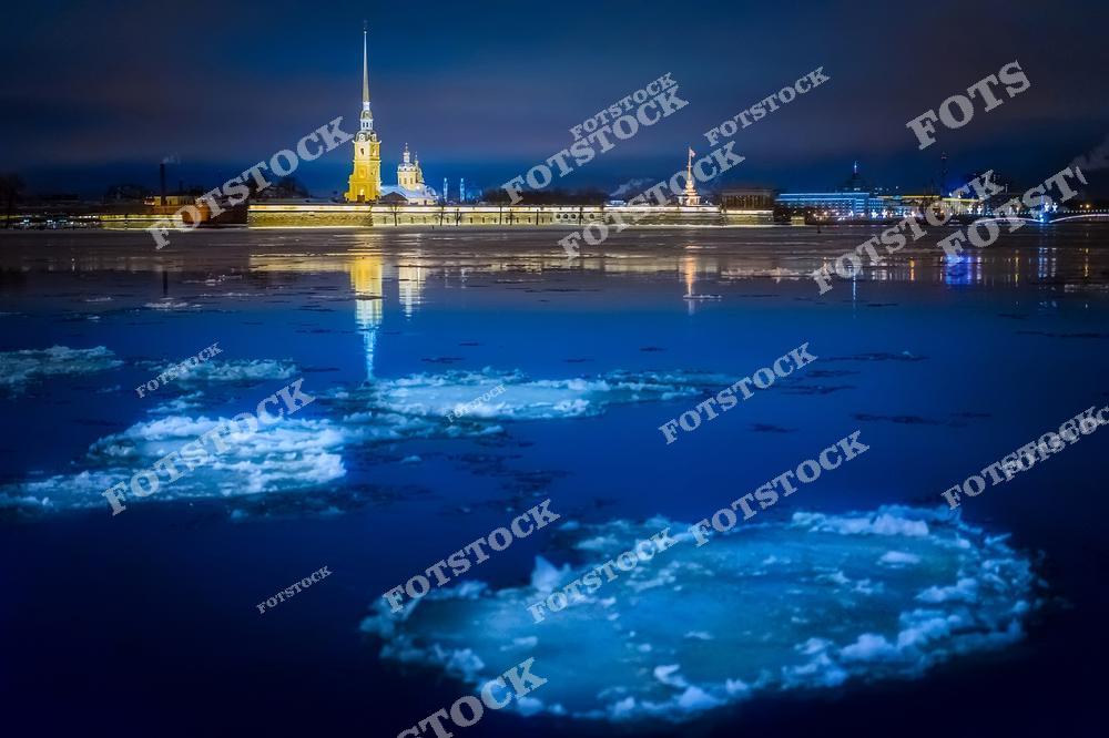 На фотографии изображена Петропавловская крепость, ярко освещенная в зимнюю ночь. На переднем плане река Нева покрыта дрейфующими льдинами, отражающими холодный свет фонарей. Высокий шпиль крепости добавляет величия этой панораме. Ночное небо и спокойствие воды создают атмосферу тишины и уюта.